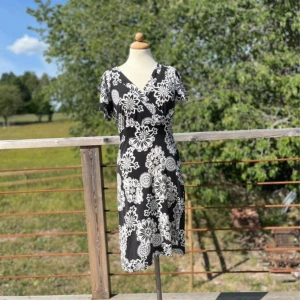 Svart blommig omlottklänning - Svart och vit omlottklänning från Ze-Ze i storlek S. Klänningen har ett stort blommigt mönster, v-ringad hals och korta ärmar. Den är knälång med en figurnära passform. Perfekt för dig som gillar en cool och feminin stil.