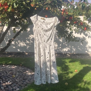 Vit blommig långklänning med dragkedja - Vit långklänning med svart blommigt mönster och korta ärmar. Klänningen har en elegant, något utställd passform och dragkedja i ryggen. Perfekt för dig som gillar stilrena och feminina plagg med en twist. Materialet känns lätt och svalt.