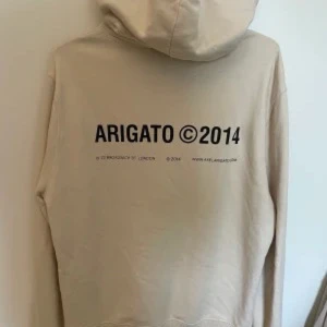 Beige hoodie från Axel Arigato - Säljer en beige hoodie från Axel Arigato med svart tryck 'ARIGATO ©2014' på bröstet och ryggen. Tröjan har huva med snörning och är tillverkad i mjuk bomull. Perfekt för en clean och stilren look.