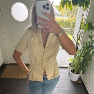 Beige kortärmad skjorta med knappar - Snygg beige skjorta med korta ärmar och klassisk krage. Skjortan har knappar framtill och två bröstfickor som ger en cool och avslappnad vibe. Perfekt att matcha med jeans för en enkel och fräsch look.