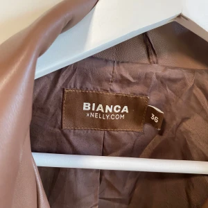 Brun skinnjacka från Bianca x Nelly - Säljer en snygg brun skinnjacka från Bianca x Nelly i storlek 36. Jackan har klassisk bikerstil med silvriga dragkedjor, tryckknappar och bred krage. Perfekt för dig som vill ha en cool och trendig look.