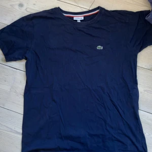 Marinblå t-shirt från Lacoste, strl 16 176cm - Snygg marinblå t-shirt från Lacoste med klassisk krokodillogga på bröstet. Enkel och stilren design med rund hals och korta ärmar. Perfekt för en clean och avslappnad look.