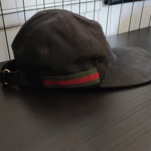 Svart keps från Gucci med GG-mönster - Svart keps från Gucci med klassiskt GG-monogram i tyget och justerbar rem i skinn baktill. Kepsen har en grön och röd rand längs sidan, typisk för Gucci. Snygg och stilren accessoar som lyfter vilken outfit som helst.