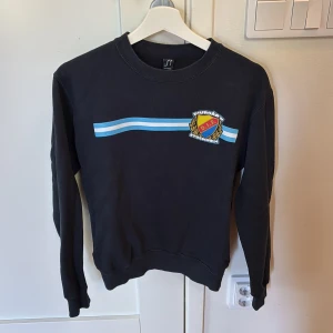 Sofialäktaren Tröja - Svart sweatshirt med Djurgårdens IF Stockholm-logga på bröstet och blåvita ränder över framsidan. Rund hals och långa ärmar, tillverkad i mjuk bomull. Perfekt för dig som vill visa ditt stöd för DIF och gillar sportig stil.
