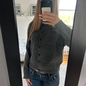 Mörkgrå kofta med knappar - Mysig mörkgrå kofta med långa ärmar och svarta knappar framtill. Tröjan är stickad och har en klassisk, enkel design som passar perfekt till jeans. Ribbstickade muddar vid ärmslut och nederkant ger en snygg finish.