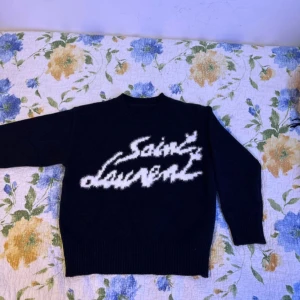 Svart stickad Saint Laurent tröja - Svart stickad tröja från Saint Laurent med vit, stor logotyp framtill i fuzzy stil. Klassisk rund halsringning och ribbade muddar vid ärmslut och nederkant. Perfekt för dig som gillar statement-plagg och exklusiv känsla. Lite small, kanske XS.