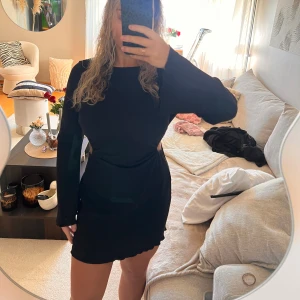 Svart ribbad kortklänning med cutout - Svart kortklänning med långärmade ärmar och snygg cutout-detalj i sidan. Klänningen är ribbad och har vågig kant nedtill för en cool touch. Materialet känns mjukt och stretchigt, perfekt för dig som gillar att sticka ut lite extra.