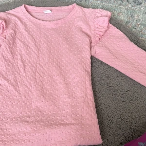 Rosa långärmad topp med volang Lindex - Söt ljusrosa långärmad topp från Lindex i storlek 146/152. Toppen har ett diskret strukturmönster och volangdetaljer på axlarna som ger en gullig touch. Perfekt för dig som gillar mjuka pasteller och fina detaljer. aldrig använt för stor för mig