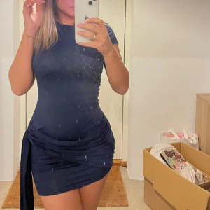 Marinblå kortklänning med knyt - Säljer en marinblå kortklänning med tight passform och snygg knyt-detalj på sidan. Klänningen har korta ärmar och är gjord i ett mjukt, stretchigt material som sitter riktigt snyggt. Perfekt för dig som gillar stilrena och trendiga plagg.