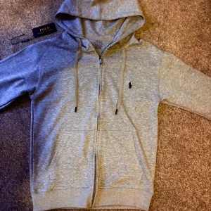 Grå hoodie från Polo Ralph Lauren - Snygg grå hoodie från Polo Ralph Lauren med dragkedja och huva. Tröjan har snörning i huvan, två fickor framtill och den klassiska broderade loggan på bröstet. Slim fit-modell i mjuk bomullsmix. Ny pris 2500kr mitt pris 699kr hör av er vid minsta fråga😁priset är inte hugget i sten 🪨 