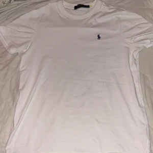 Vit t-shirt från Polo Ralph Lauren - Vit klassisk t-shirt från Polo Ralph Lauren i bomull. T-shirten har korta ärmar, rund halsringning och en liten mörkblå broderad logga på bröstet. Enkel och stilren modell som passar till allt.
