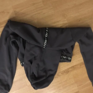 Mörkgrå croppad hoodie från ICIW - Säljer en mörkgrå croppad hoodie från ICIW med långa ärmar och huva. Tröjan har svarta detaljer med ICIW-logga längs nederkanten och ärmarna. Perfekt för en sportig och avslappnad stil.