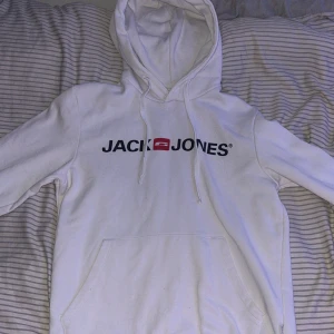 Vit hoodie från Jack & Jones - Säljer en stilren vit hoodie från Jack & Jones med svart och rött logotryck på bröstet. Tröjan har huva med snörning, stor magficka och är tillverkad i mjuk bomullsmix. Perfekt för dig som gillar en clean och enkel look. Perfekt att ha inomhus och ute den värmer dig 