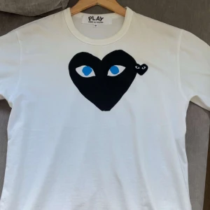 Vit Comme des Garçons Play t-shirt - Snygg vit t-shirt från Comme des Garçons Play med det ikoniska svarta hjärtat med ögon och en mindre hjärtdetalj på bröstet. Klassisk rund hals och korta ärmar. Perfekt för dig som gillar streetstyle och vill sticka ut.