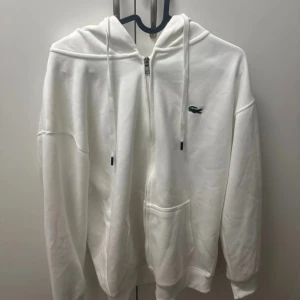 Vit hoodie med dragkedja från Lacoste - Vit hoodie från Lacoste i regular fit med dragkedja framtill. Mjuk bomullskänsla och känguruficka gör den både stilren och chill. Perfekt för dig som gillar cleana och sportiga vibes.