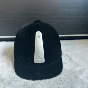 Svart sammet ridhjälm från KYLIN - Snygg svart ridhjälm från KYLIN i mjuk sammet med silverfärgad metallplatta framtill. Klassisk rundad form med kort skärm och ventiler på sidorna. Perfekt för dig som vill ha en stilren look när du rider.