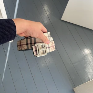 Burberry rutigt bälte med B-spänne - Säljer ett snyggt Burberry bälte med klassiskt rutigt mönster i beige, svart och grå toner. Bältet har ett stort metallspänne med ett B i silverfärg. Materialet är slitstarkt och har en lyxig känsla, perfekt för att lyfta din outfit.