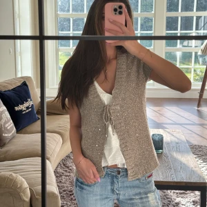 Beige stickad väst/kofta med paljetter - Säljer en beige stickad väst/kofta med korta ärmar och v-ringning. Koftan har en snygg knytning framtill och är dekorerad med små paljetter som ger en glittrig look. Perfekt att slänga över en topp💕