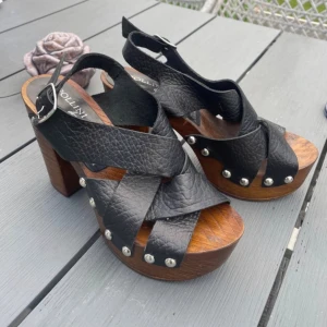 Pollini svarta sandaler med träklack - Snygga sandaler från Pollini med breda svarta läderremmar och silverfärgade nitar. Skorna har en rejäl träklack och plattform, samt öppen tå och justerbart spänne vid ankeln. Perfekta för dig som gillar statementskor med retrovibbar. Köpta för 1000kr+