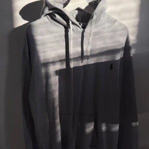 Grå hoodie från Ralph Lauren - Säljer en stilren grå hoodie från Ralph Lauren med klassisk huva och snörning. Tröjan har en broderad logga på bröstet och en stor ficka framtill. Perfekt för en chill och avslappnad look.