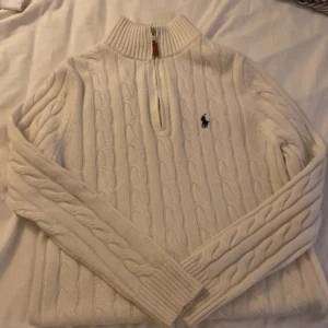 Använd två gånger Vit kabelstickad tröja Polo Ralph Lauren - Kabelstickad vit tröja från Polo Ralph Lauren med halv dragkedja och ribbad hög krage. Klassisk broderad logga på bröstet och snygga detaljer på dragkedjan. Perfekt för lager-på-lager och chill höst- och vinterstil.