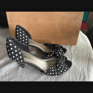 Svarta prickiga klackar - Säljer ett par svarta pumps med vita prickar från VOX Shoes. Skorna har öppen tå, låg klack och en snygg rosettdetalj framtill. Perfekta för dig som gillar retrostil och vill sticka ut med ett lekfullt mönster.