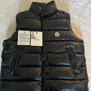 Svart Moncler vest 🔥🔥🙌🙌 - Snygg svart dunväst från Moncler med glansig finish och klassisk logga på bröstet. Västen har hög krage, två dragkedjefickor framtill och quiltad design. Perfekt att slänga över en hoodie eller tröja för extra värme. Insidan har en cool serietidningsdetalj. Pris kan diskuteras 🙌🙌🔥