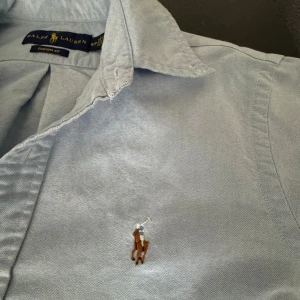 Ljusblå skjorta från Ralph Lauren - Klassisk ljusblå skjorta från Ralph Lauren med broderad polospelare på bröstet. Skjortan har knappar framtill, klassisk krage och är i bomull. Perfekt för dig som gillar stilren och tidlös design. ( En knapp från höger armen är trasig men man märker inte så mycket ).