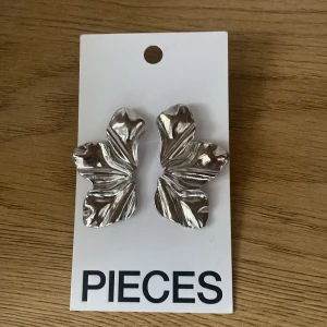 Silvriga bladmönstrade örhängen Pieces - Stora statementörhängen från Pieces i blankt silvrigt material, formade som stiliserade blad med veckad yta. Perfekta för dig som vill sticka ut med en cool och trendig accessoar. Säljs i originalförpackning och passar till alla outfits.