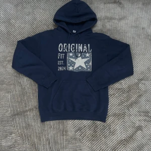 Scandi hoddie  - Mörkblå hoodie från Original Fit med stort tryck framtill som har stjärnor och texten 'Original Fit Est. 2024'. Tröjan har huva med snörning, magficka och långa ärmar. Perfekt för chill dagar eller när du vill ha en avslappnad look.