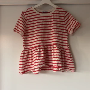 Randig volangblus från Zara - Superfin blus från Zara med röda och vita ränder. Modellen har korta ärmar, rund halsringning och en volangkant nedtill som ger en söt och avslappnad look. Tillverkad i mjuk bomull, perfekt för vår och sommar.