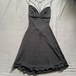 Svart kort klänning från Oh Polly - Supersnygg svart kort klänning från Oh Polly med tunna axelband och öppen rygg med snörningsdetaljer. Klänningen har en lätt glansig finish och figurnära passform upptill som övergår i en lätt utsvängd kjol. ( helt oanvänd)