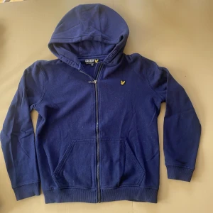 Marinblå hoodie från Lyle & Scott - Snygg marinblå hoodie från Lyle & Scott med dragkedja och huva. Klassisk gul logga på bröstet och två fickor framtill. Perfekt för dig som gillar stilrena och bekväma plagg.
