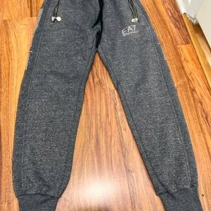 Mörkgrå EA7 mjukisbyxor med dragkedjor - Mörkgrå mjukisbyxor från EA7 Emporio Armani med svarta snören i midjan, ribbade muddar vid benslut och två fickor med dragkedjor. Snygga detaljer med EA7-logga på ena benet och diskreta ränder längs sidorna. Perfekta för chill eller träning.
