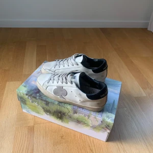 Vita sneakers från The Gallerist - Snygga vita sneakers från The Gallerist med svart häl och grå klöverdetalj på sidan. Skorna har klassisk snörning, platt sula och rund tå. Tillverkade i skinn med detaljer i mocka. Perfekta för dig som gillar stilrena skor. Otroligt skön och mjuk sula. 