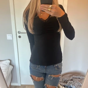 Långärmad topp - Svart långärmad topp från Anna Field med v-ringning och spetsdetalj vid halsen. Toppen har rynk framtill och är figurnära i modellen. Funkar både i vardag och till fest❤️Storleken är XS-S