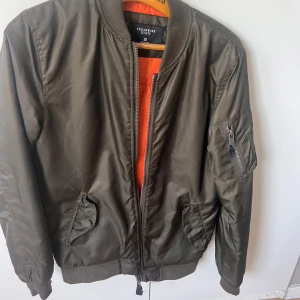 Olivgrön bomberjacka Cedarwood State XS - Snygg olivgrön bomberjacka från Cedarwood State i storlek XS. Jackan har orange foder, klassisk ribbad krage och muddar, samt dragkedja framtill och på ärmen. Tillverkad i syntetmaterial och har två fickor framtill.