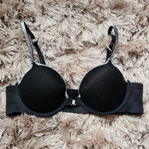 Svart bh med silverdetaljer 80B - Svart bh med vadderade kupor och diskret rutmönster. Axelbanden har silverfärgade kanter och justerbar längd. Bh:n har en smidig metallknäppning fram och är tillverkad i mjuk polyamid och elastan för skön passform.