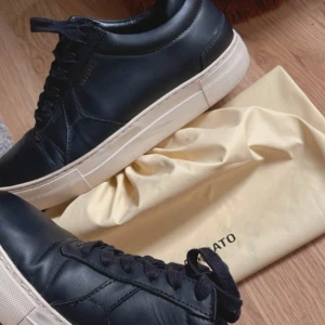 Svarta sneakers från Axel Arigato - Snygga svarta sneakers från Axel Arigato med vit platt sula och diskret logga på sidan. Skorna är i skinn och har klassisk snörning. Perfekta för dig som gillar stilrena och minimalistiska sneakers med premiumkänsla.