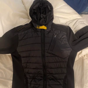Svart Parajumper cardigan - Nästan aldrig använd. Storlek M men passar Även S. Säljer pga ska köpa ny jacka. (OBS köpte för 3 tusen på plick) svart cardigan från Parajumpers med huva, gul detalj vid halsen och orangea inslag på insidan. Jackan har quiltad framsida och rygg, dragkedja framtill och bröstficka med dragkedja. Märkespatch på ena ärmen. Perfekt för kyliga dagar.