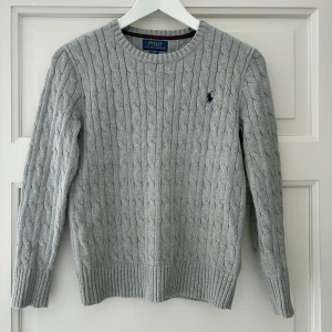 Grå stickad tröja från Polo Ralph Lauren för kille - Snygg grå kabelstickad tröja från Polo Ralph Lauren för kille. Storlek M (10-12) står L i nacken men måste vara felmärkt. Knappt använd i mycket fint skick. 