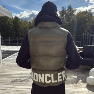 Grön dunväst från Moncler - Snygg dunväst från Moncler i olivgrönt med stor vit logga baktill. Västen har en hög krage, dragkedja framtill och insida i orange. Perfekt för lager-på-lager och streetwear-vibe.