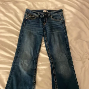 Blå bootcut jeans med fickdetaljer Lågmidjade - Snygga blå jeans med bootcut-modell och coola slitningar framtill. Byxorna har klassiska fem fickor och dekorativa knappar på bakfickorna. Perfekta för dig som gillar en avslappnad men trendig look.