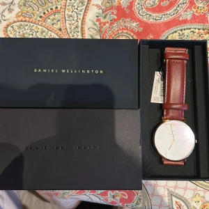 Daniel Wellington klocka med brunt läderband - Stilren klocka från Daniel Wellington med vitt ur och brunt läderarmband. Urtavlan är rund och minimalistisk med tunna guldiga markeringar och visare. Klockan har klassisk design och levereras i originalask. Helt ny och inte använd. Nya priset på Boozt 1699. 