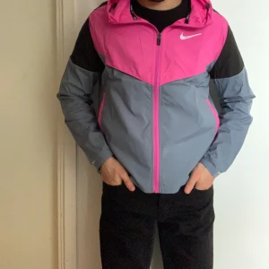 Nike jacka i rosa - Nike vindjacka i fräsch rosa och svart färg med grå detaljer. Jackan har huva, hel dragkedja och två fickor med dragkedja. Swoosh i silver på bröstet. Perfekt för blåsiga dagar och sportiga outfits. Obs jackan är I Dri-fit sitter tajt och fint på kroppen !