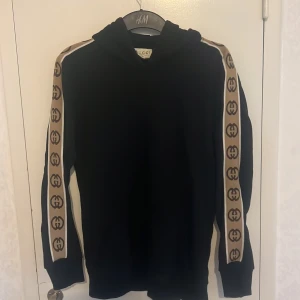Svart Gucci hoodie med logoband - Svart hoodie från Gucci med huva och beige logoband längs ärmarna. Ikoniska GG-loggor i brunt på banden ger en lyxig vibe. Tillverkad i mjuk bomull för maximal komfort. Perfekt för dig som vill sticka ut med en stilren och exklusiv look.