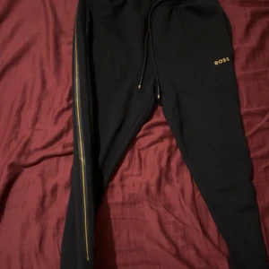 Svarta mjukisbyxor från BOSS med gulddetaljer - Svarta mjukisbyxor från BOSS med guldfärgad logga på låret och guldfärgad rand längs ena benet. Byxorna har snörning i midjan och är tillverkade i mjukt material för maximal komfort. Perfekta för en chill och stilren look. Nytt pris 1400 men säljes för 420kr. Kvitto finns och påse från affären Room 1006