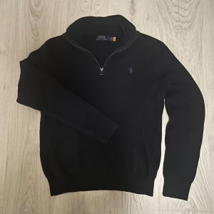 Ralph lauren tröja  - Mörkblå stickad tröja från Polo Ralph Lauren med halv dragkedja och klassisk broderad logga på bröstet. Tröjan har lång ärm, ribbade muddar och en normal passform. Perfekt till jeans för en clean och stilren look.