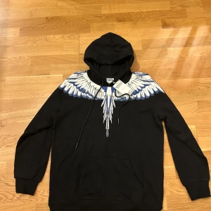 Marcelo Burlon svart hoodie med vingar - Svart hoodie från Marcelo Burlon med ikoniskt vingtryck i vitt och blått över axlar och bröst. Tröjan har huva med snörning och känguruficka framtill. Tillverkad i mjuk bomull, perfekt för dig som vill sticka ut med en unik streetstyle.