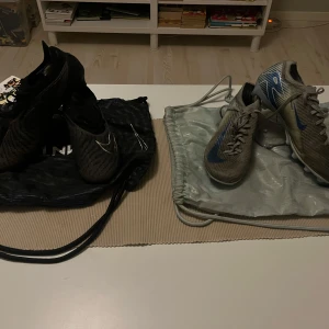 Nike Air Zoom fotbollsskor grå & svarta - Två par Nike Air Zoom fotbollsskor säljes! Ena paret är gråa med blå detaljer och snörning, det andra är svarta med silverdetaljer och mönstrad ovandel. Båda har platt sula med dobbar och är tillverkade i syntetmaterial. Perfekta för dig som vill sticka ut på planen.